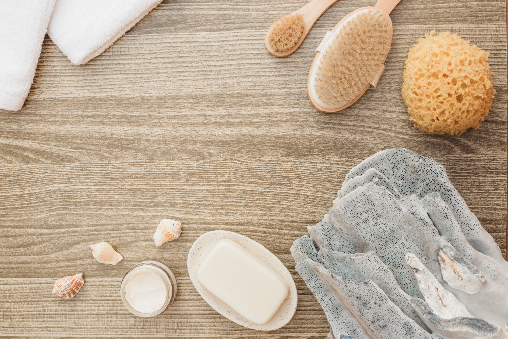 Spa and wellness setup featuring a white towel, natural bristle body brush, sea sponge, bar of soap, open jar of cream, and seashells on a wooden textured background.