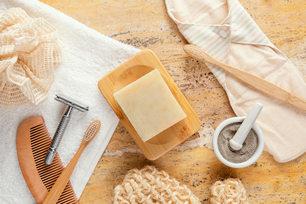 Zero waste bathroom accessories on a yellow textured background, including a white towel, wooden soap dish with bar soap, bamboo toothbrushes, body brush, loofah, safety razor, and a mortar with grey powder.