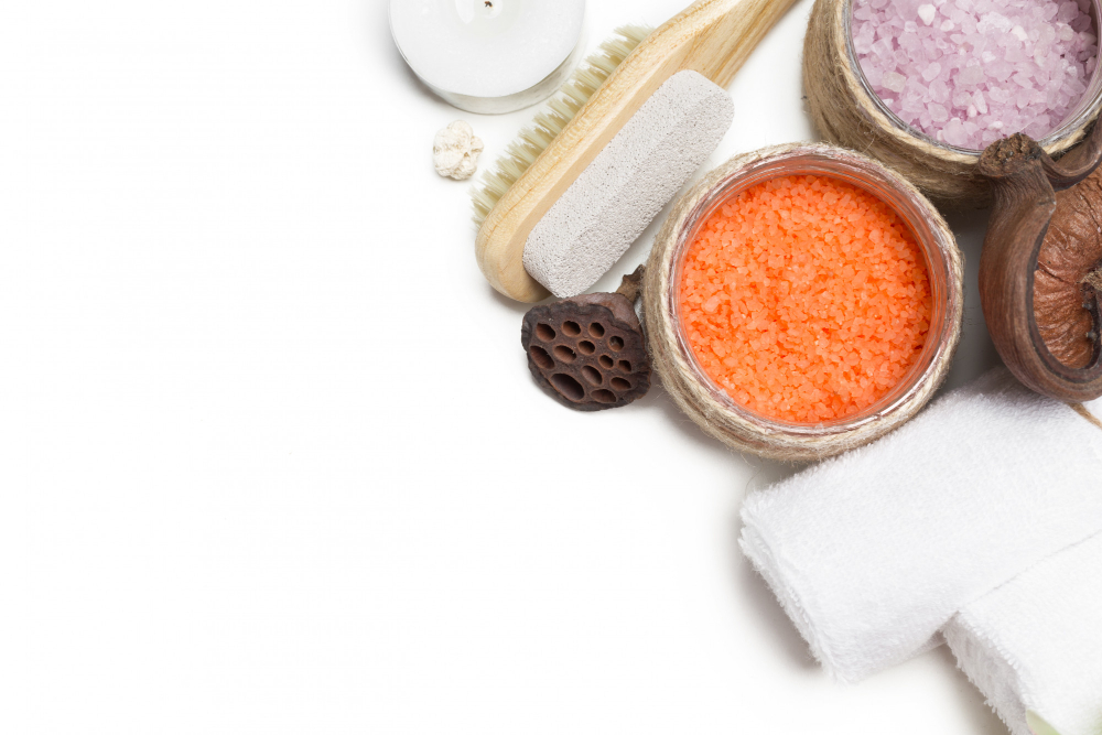 Spa essentials with bath salts in wooden bowls, a white candle, towel, and natural brushes on a white background.